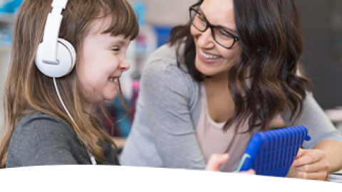 A female elementary student wearing headphones smiles while working with her teacher on a classroom digital tablet.